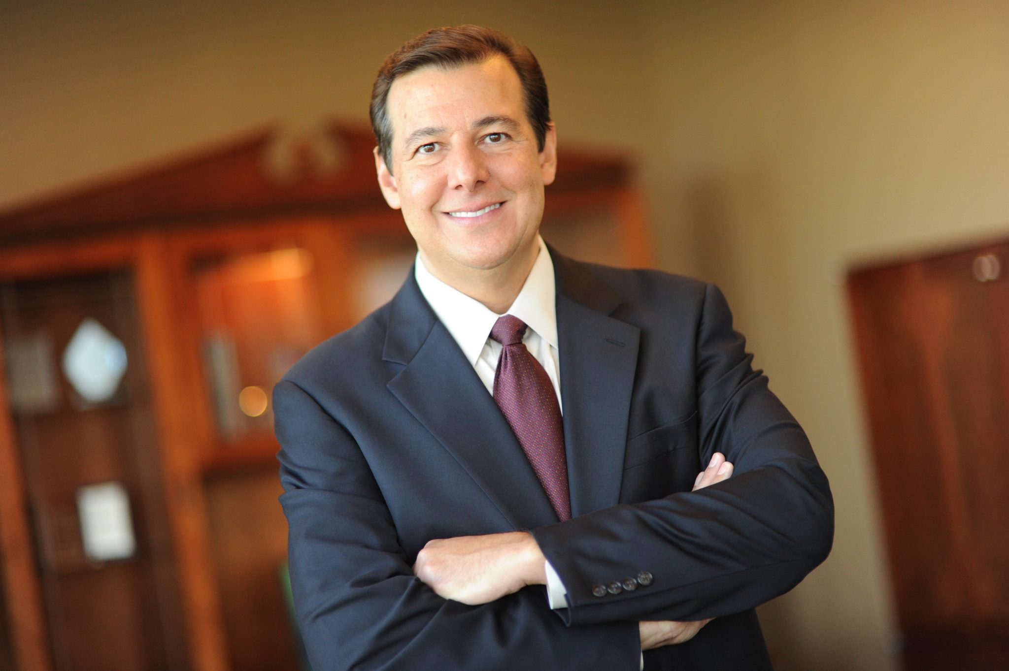 A.J. Kazimi, Chief Executive Officer, smiling with arms crossed in a suit, standing in an office setting.